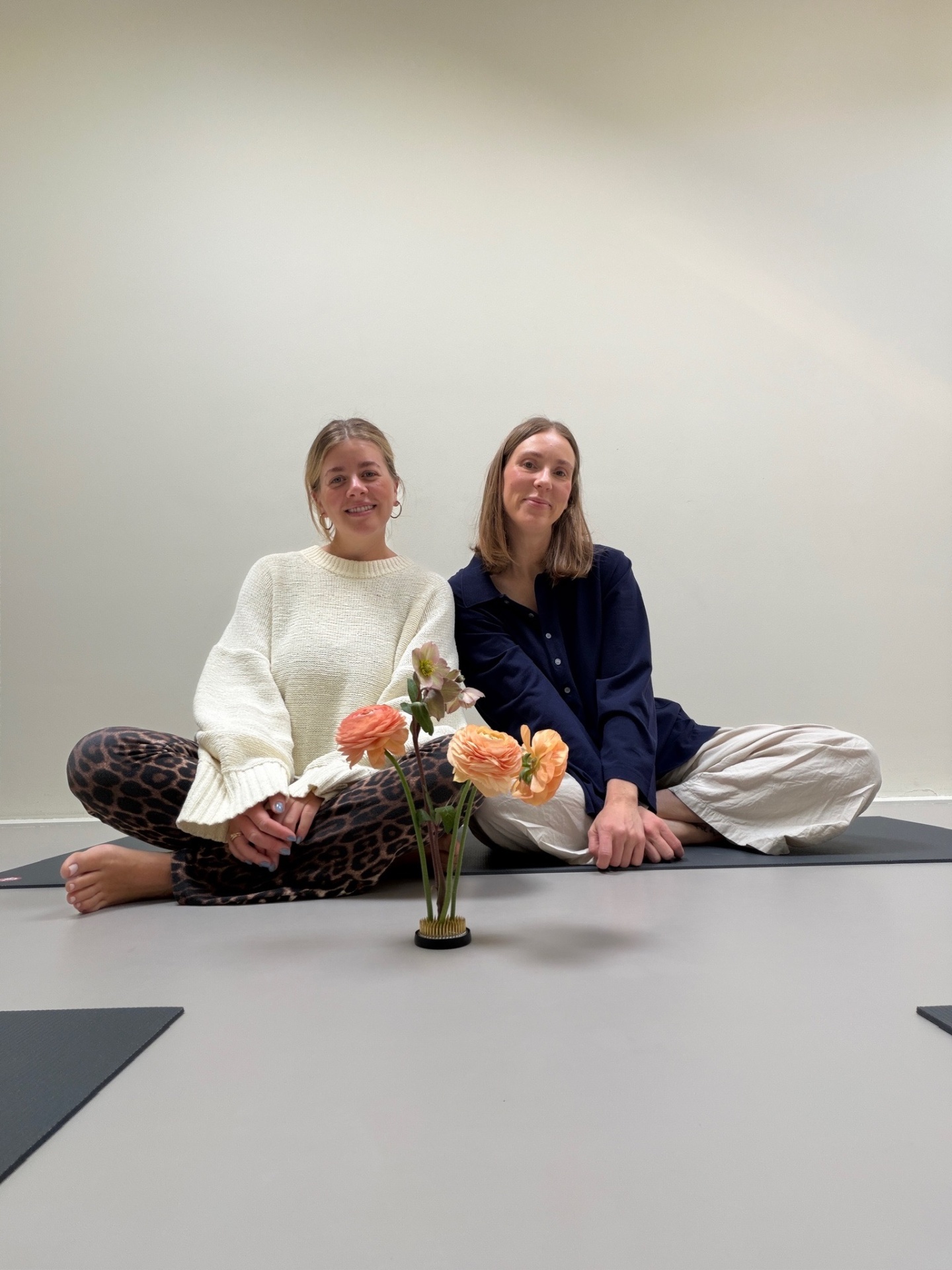 Kat and Jaimee, founders of The Flowr House, sitting on yoga mats with a flower arrangement